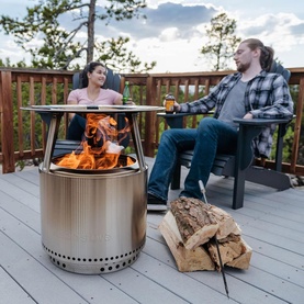 Solo Stove Bonfire Heat Deflector in stainless steel, shown in use on a deck with two people relaxing around an open fire pit