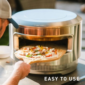 Compact countertop pizza oven in pale blue and steel, with a freshly topped pizza being placed inside on a wooden peel outdoors.