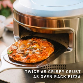 Stainless steel countertop pizza oven with cooked mushroom pizza on peel, promotional text overlay reads Twice As Crispy Crust As Oven Rack Pizza