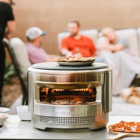 Solo Stove Pi Prime outdoor pizza oven in brushed stainless steel, shown in use with pizza cooking inside, friends gathered in background