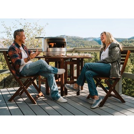 Two people seated on a wooden deck with a stainless steel outdoor pizza oven on the table, set against a scenic mountain backdrop.