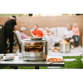 Stainless steel outdoor pizza oven with glowing flames, on a table with pizza slices, family relaxing in garden background.