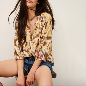 Model wearing Bash yellow abstract ibukat-print blouse with tie neck and blouson sleeves, styled with denim shorts and layered necklaces