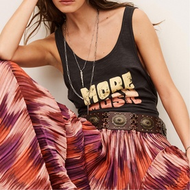 Model wearing Bash Womens French Dress in orange pleated midi skirt with black graphic tank top, brown studded belt and layered necklaces.