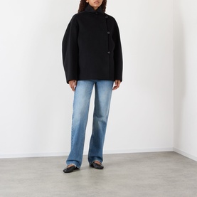 Model wearing Rohe Double-Faced Asymmetric Jacket in black with funnel neck and off-centre buttons, styled with blue jeans and black flats.