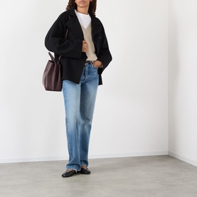 Model wearing Rohe Double-Faced Asymmetric Jacket in black with blue straight-leg jeans, white top, and black loafers, carrying a dark burgundy bag.