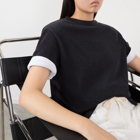 Model wearing Rohe Anthracite Melange merino wool T-shirt with white contrast cuffs, styled with cream wide-leg trousers, seated on a chair.