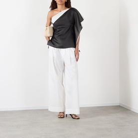 Model wearing Rohe black silk one-shoulder top with draped detail, styled with white wide-leg trousers and a cream bag.