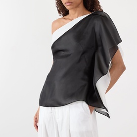 Model wearing Rohe black silk one-shoulder top with draped cape detail, styled with white wide-leg trousers