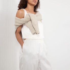 Model wearing Rohe cream silk trousers with white vest top and beige knit cardigan draped over shoulders