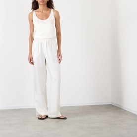 Model wearing Rohe cream silk wide-leg trousers with elasticated waist, styled with a white vest top and brown flat sandals.