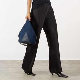 Model carrying Stella McCartney Appaloosa Bucket Bag in navy faux suede with gold-tone hardware, styled with black wide-leg trousers and heels.