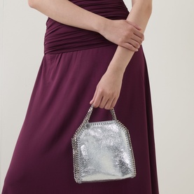 Model carrying Stella McCartney Falabella Mini Crossbody Bag in silver metallic finish with chain trim and top handle, styled with a plum dress.