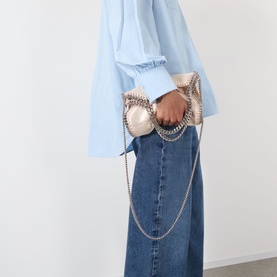Model carrying a metallic silver fold-over clutch bag with silver chain detailing, styled with a light blue blouse and denim midi skirt.