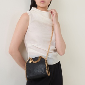 Model carrying Stella McCartney Falabella Tiny Tote Bag in black with gold-tone chain trim, carry handles, and crossbody chain strap. Styled with