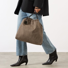 Model carrying Stella McCartney Falabella Medium Drawstring Tote Bag in dark taupe with chain handles, styled with black blazer and jeans