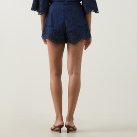 Model wearing Zimmermann blue cotton shorts with scalloped broderie anglaise hem, styled with matching top and tan mules, rear view