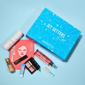 Selection of beauty products on a blue background including an eyeshadow palette, setting spray, facial gel, and shimmer mist