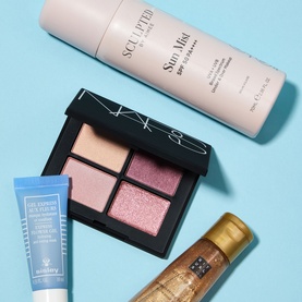 Beauty gift set featuring eyeshadow palette, face mist, facial gel, and shimmer body oil arranged on a blue background.