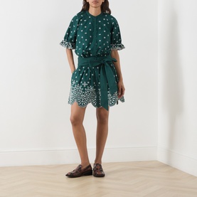 Model wearing Essentiel Antwerp bottle green cotton mini dress with floral broderie anglaise, belted waist and short sleeves, styled with loafers.