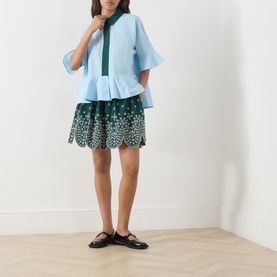 Model wearing Essentiel Antwerp aquamarine cotton shirt with flutter sleeves and peplum hem, styled with dark green embroidered mini skirt and black