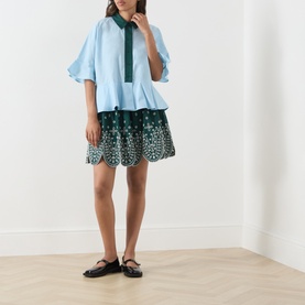 Model wearing Essentiel Antwerp bottle green cotton lace scallop-hem mini skirt, styled with a light blue ruffle blouse and black ballet flats.