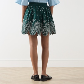 Model wearing Essentiel Antwerp bottle green cotton mini skirt with scalloped broderie anglaise hem, styled with black loafers, rear view.