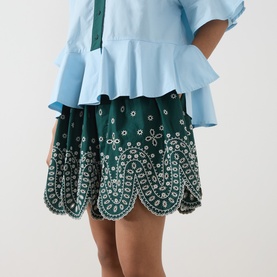 Model wearing Essentiel Antwerp bottle green cotton mini skirt with floral eyelet embroidery and scalloped hem, styled with a light blue ruffled top.