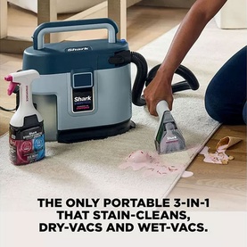 Shark EveryMess Portable 3-in-1 Stain Eliminator being used on a carpet spill, with cleaning spray bottle alongside, in blue-grey.