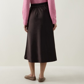 Model wearing InWear Chocolate Brown satin midi skirt, rear view, styled with a pink knit cardigan and tan flat shoes.