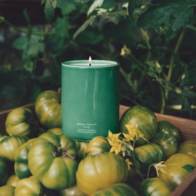 Jo Malone Green Tomato Vine Candle in green ceramic vessel, lit, surrounded by green tomatoes and foliage