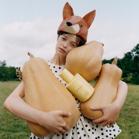 Model with Jo Malone Velvety Butternut Cologne, wearing fox hat and polka dot dress, holding oversized butternut squashes outdoors.