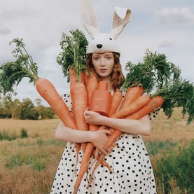 Jo Malone Carrot Blossom Cologne lifestyle shot featuring model in polka-dot dress and bunny hat holding fresh carrots in a field