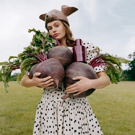 Jo Malone Scarlet Beetroot Cologne bottle held among large fresh beetroots by model in polka dot dress and hat, outdoor field setting