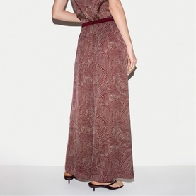 Model wearing Jigsaw red abstract print maxi dress with velvet belt detail, shown from behind, styled with black heeled mules