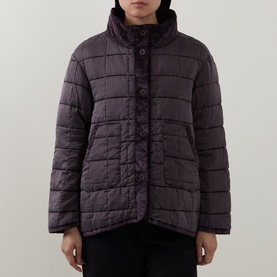Model wearing Transit quilted jacket in Choco, featuring horizontal quilting, high collar, snap button fastening and patch pockets.