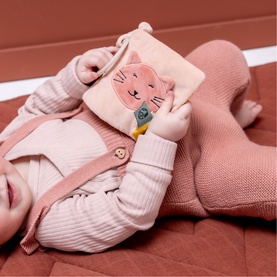 Baby holding Trixie pink soft fabric cat activity book, lying on rose-toned knit blanket, dressed in pink striped outfit