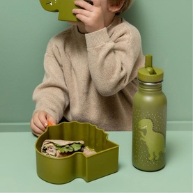 Child using Trixie green dinosaur-shaped lunch box with matching green water bottle with straw on a green surface