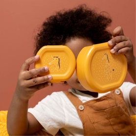 Young child holding two Trixie orange lunch boxes up to their face against a terracotta background.