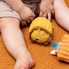 Trixie yellow lion soft rubber ball toy shown with baby on orange textured blanket, with matching lion finger puppet