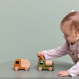 Toddler playing with Trixie wooden toy trucks in tan and green, on a grey surface against a sage green background
