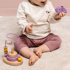 Trixie wooden activity toy set in pastel purple and natural wood tones, shown with baby sitting on floor holding a puzzle piece
