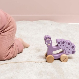 Trixie purple wooden peacock push-along toy on wheels, shown on cream rug with baby in background