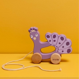 Trixie wooden pull-along peacock toy in purple with decorative tail, rolling wheels, and cotton pull string, on yellow background.