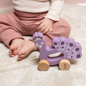 Trixie purple wooden peacock pull-along toy on wheels, shown with seated baby in background