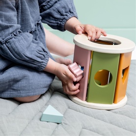 Trixie multicolour wooden shape sorter toy with pastel pink, green and orange panels, shown with child's hands placing shapes
