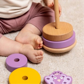 Trixie wooden stacking ring toy in purple, yellow and natural wood tones, shown with baby playing on grey floor