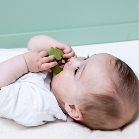 Baby lying on back holding and mouthing a Trixie green turtle-shaped toy