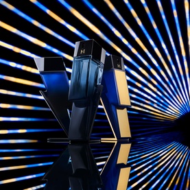 Carolina Herrera Bad Boy EDP fragrance bottles in navy blue and gold, displayed against a dramatic blue and gold ray backdrop with mirror reflection