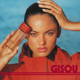 Model with Gisou Body Mist held up to face, wearing red jacket, against blue background